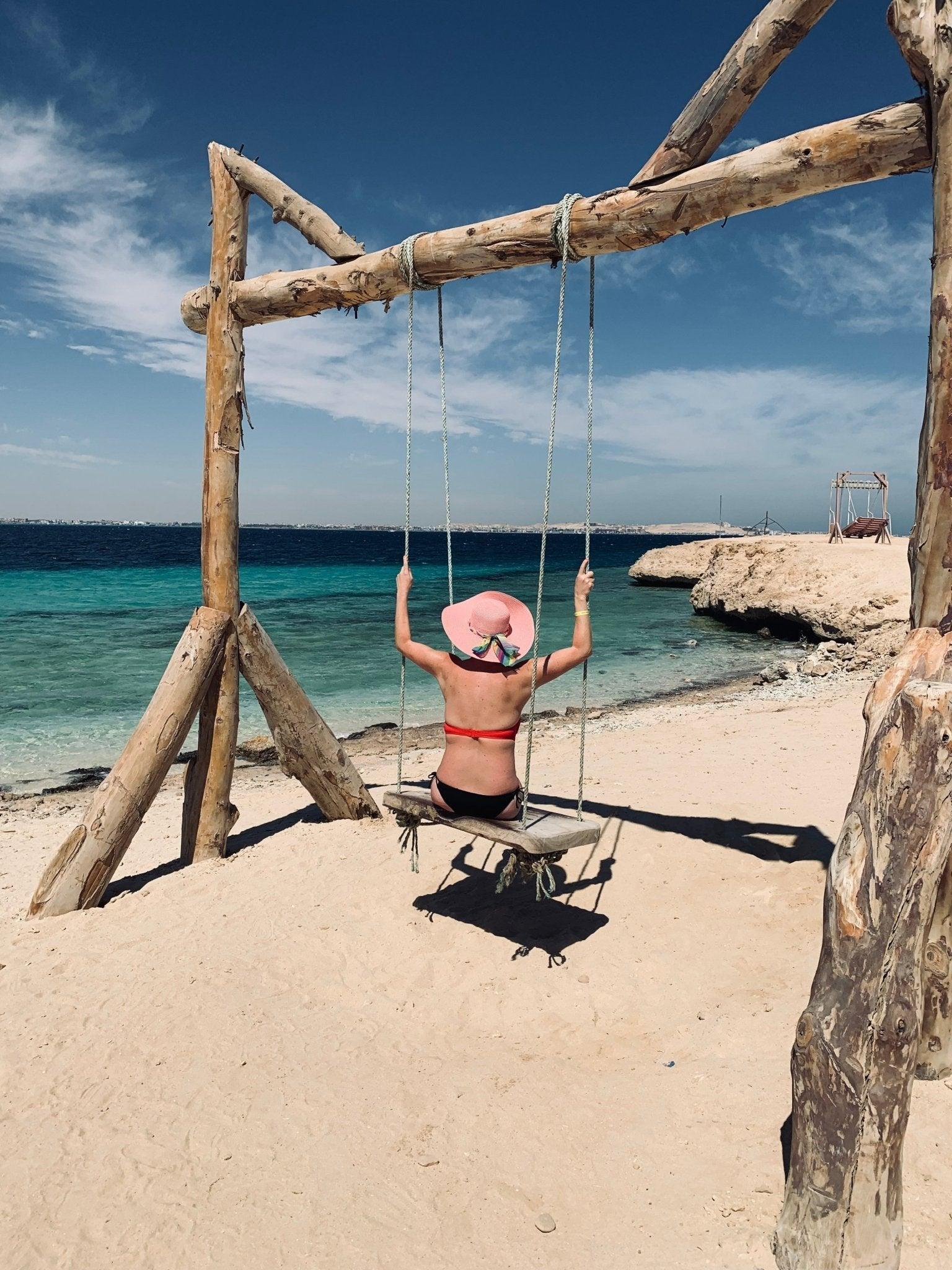 Frau im Bikini und rosa Hut auf Holzschaukel am Strand mit türkisem Meer und blauem Himmel